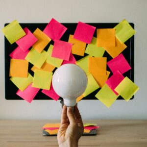 Hand holding a light bulb in front of colorful sticky notes on monitor, symbolizing brainstorming and innovation.